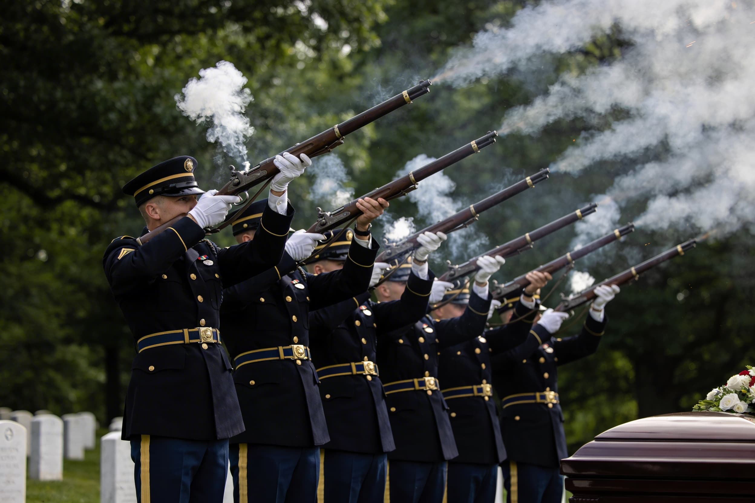 A rifle team of seven service members in dress uniforms firing a three-volley salute at a military cemetery graveside ceremony