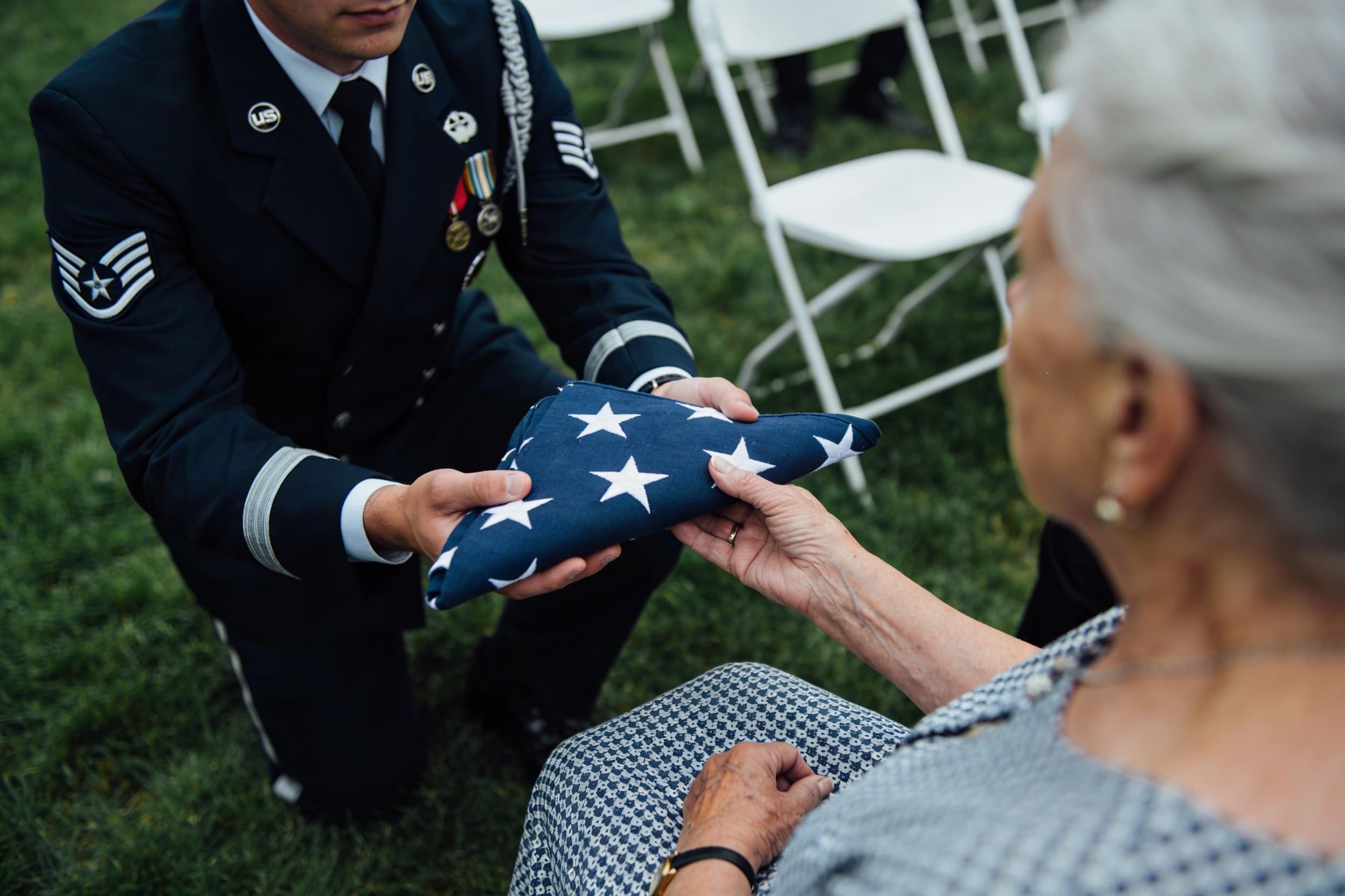 A uniformed service member kneeling to present a triangular folded American flag to a seated family member at a graveside military funeral