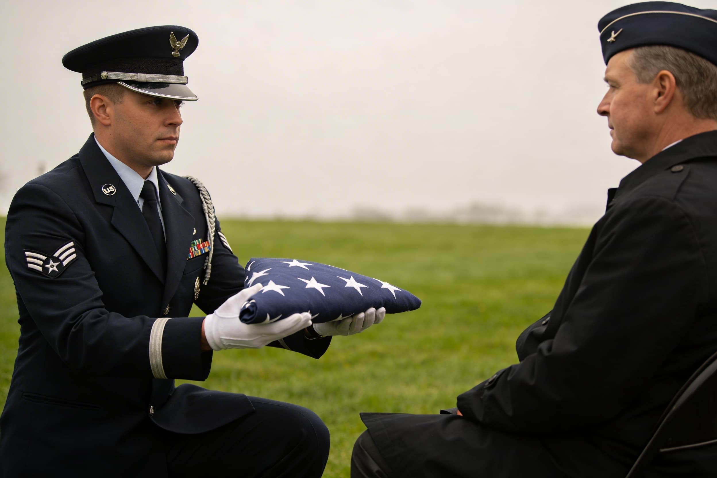 A military honor guard member in dress uniform presenting a tightly folded triangular American flag to a seated family member