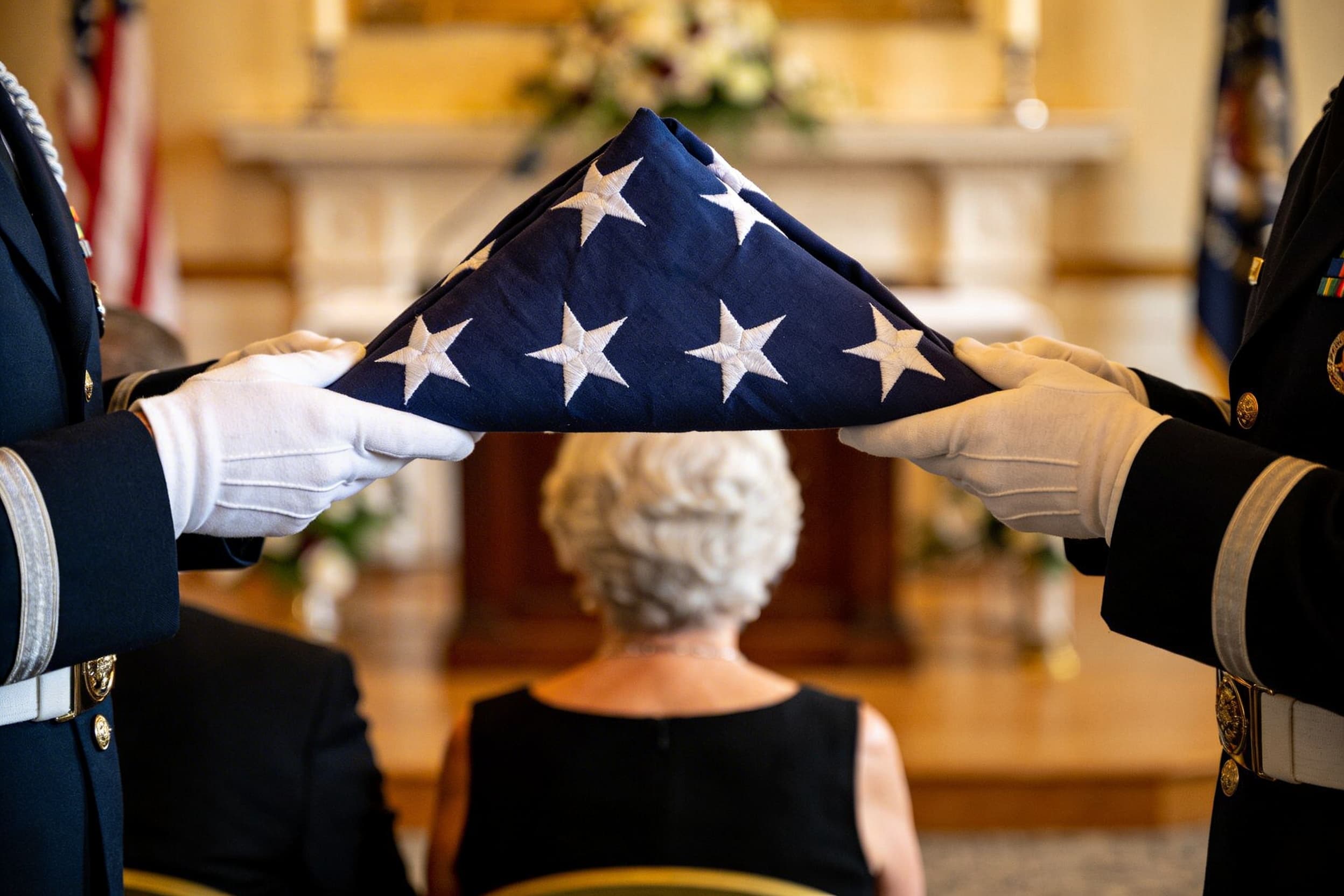 A folded triangular American flag held at waist height by gloved hands in military dress uniform, presenting to a seated family member, dignified indoor setting
