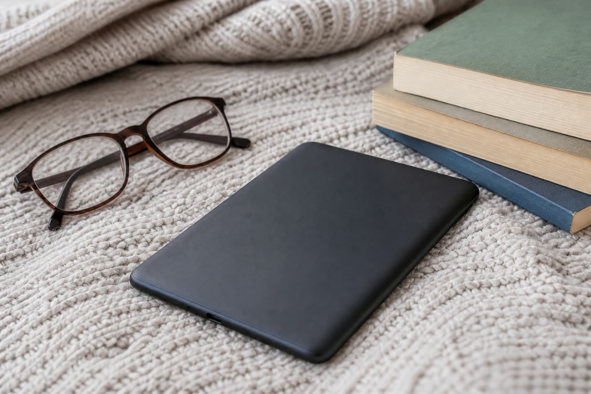 An e-reader tablet lying face-down on a knitted blanket beside a pair of reading glasses and a stack of paperback books