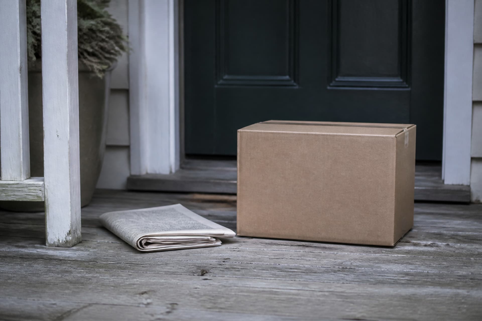 A single unopened cardboard delivery box resting on a doorstep beside a folded newspaper, flat morning light