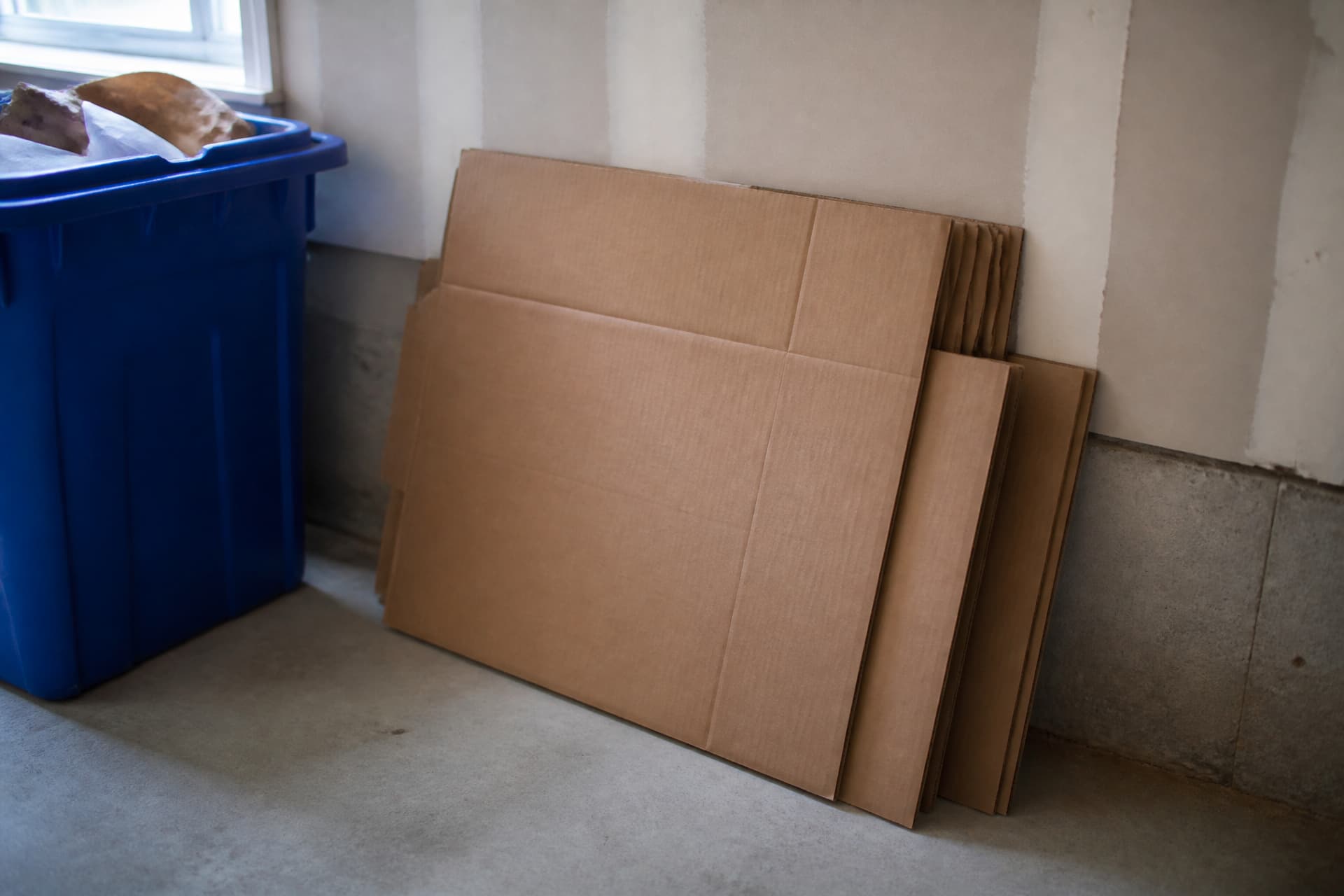 Three flattened cardboard delivery boxes stacked against a wall beside a recycling bin in a garage, cool light from a window