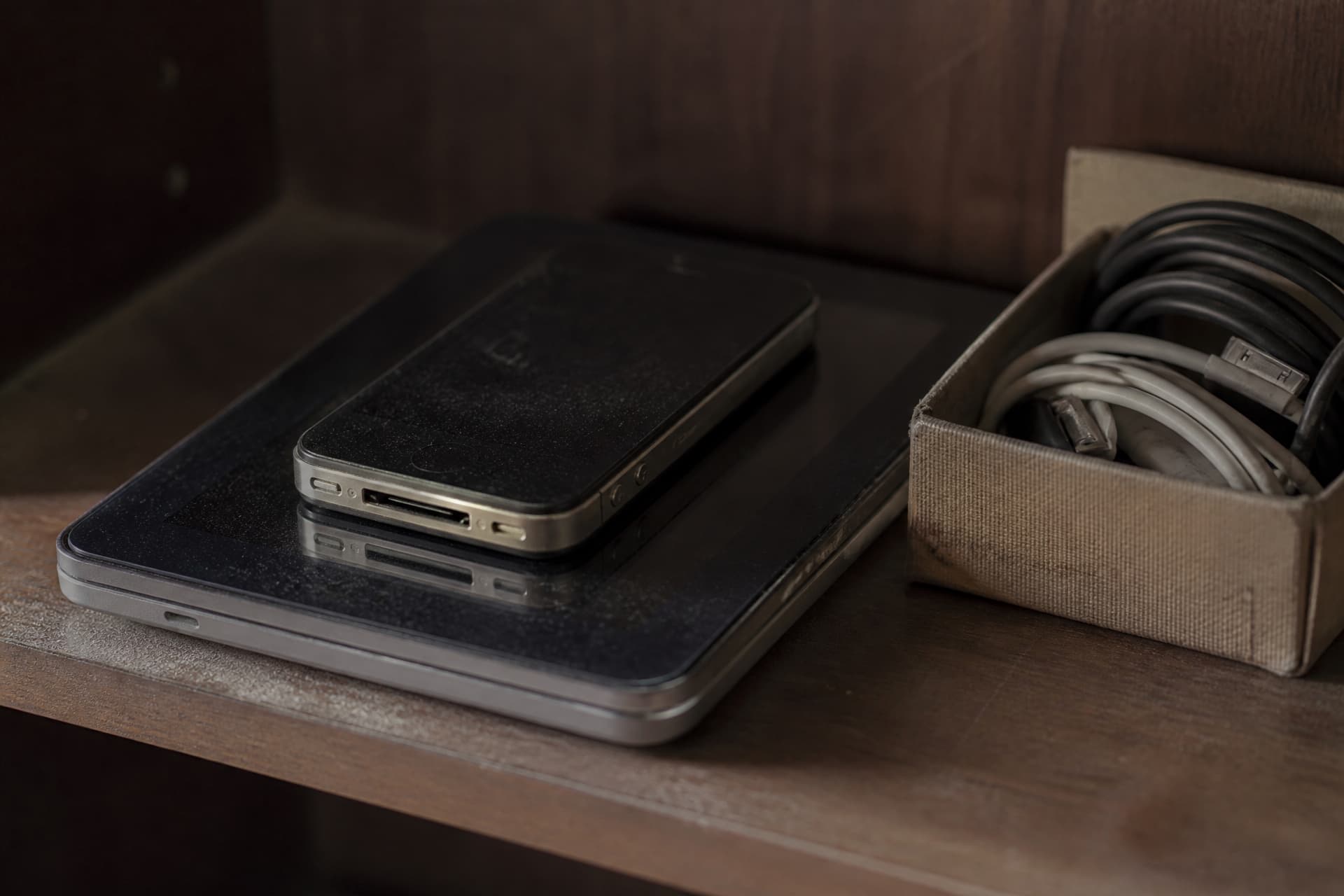 An older phone and a tablet stacked on a shelf beside a small box of cables, dust visible in the light