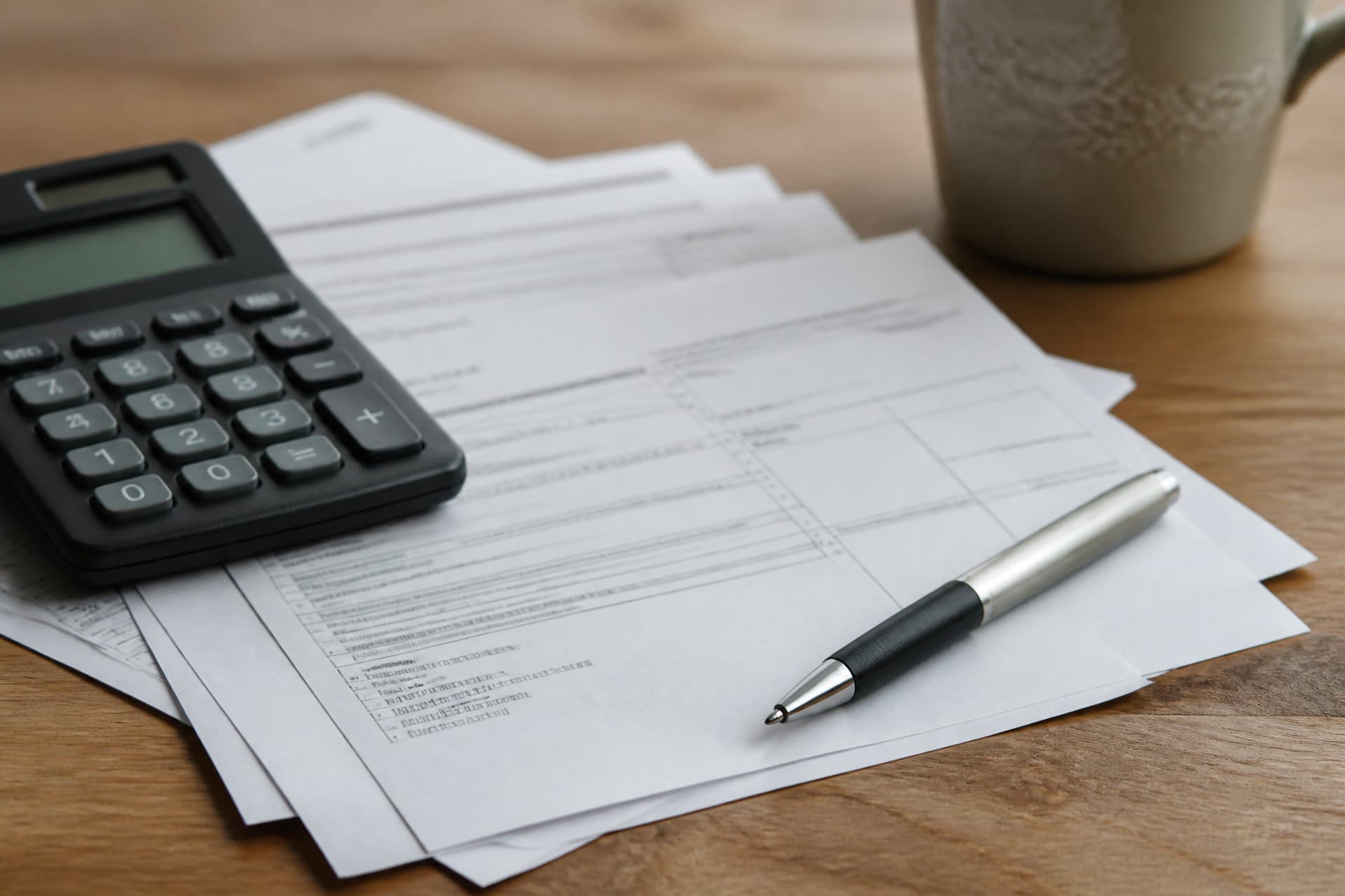 A calculator and a pen resting on a small stack of printed statements on a wooden desk, flat afternoon light