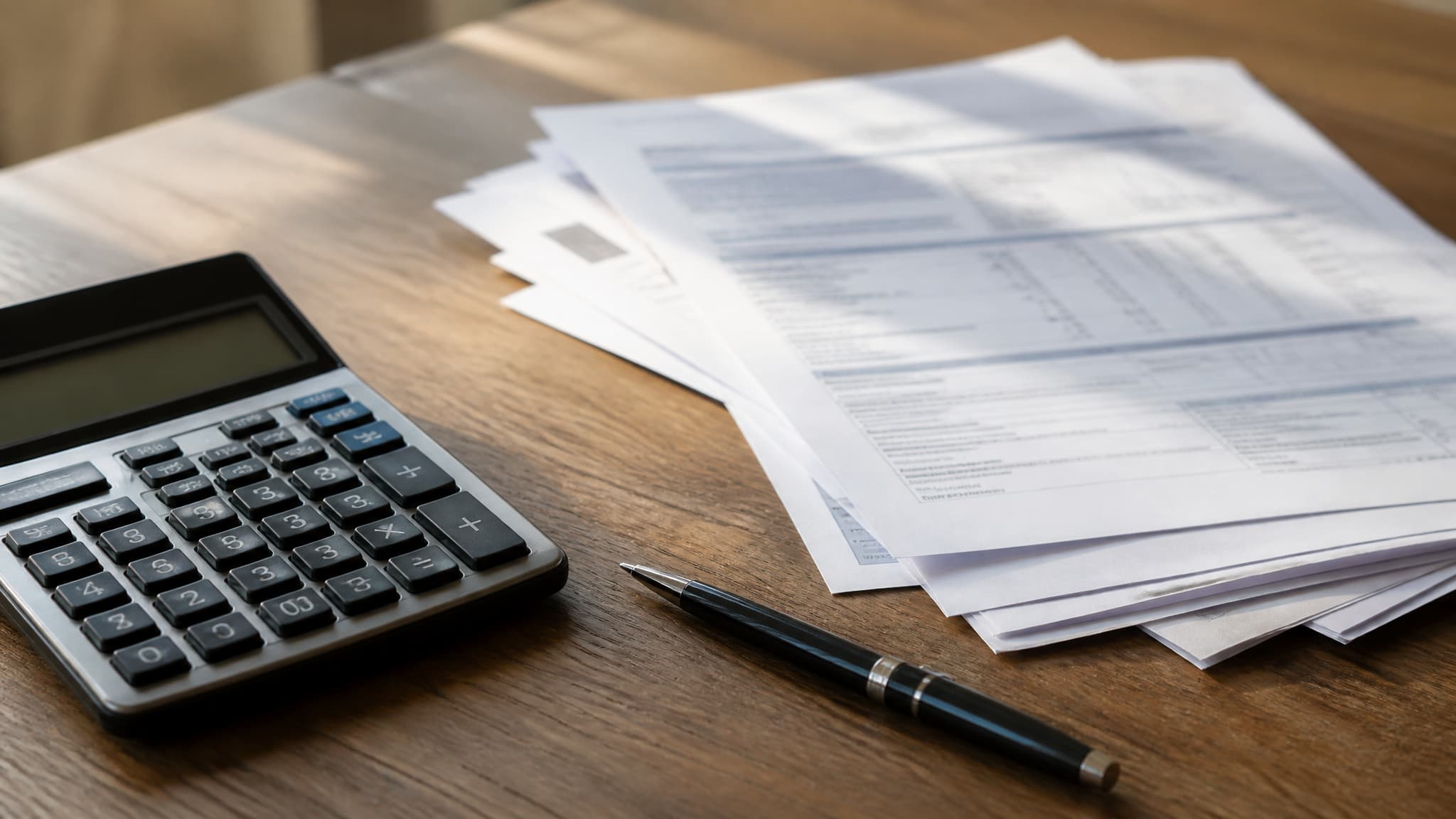 A calculator, a pen and a small stack of printed statements on a wooden desk, in flat afternoon light