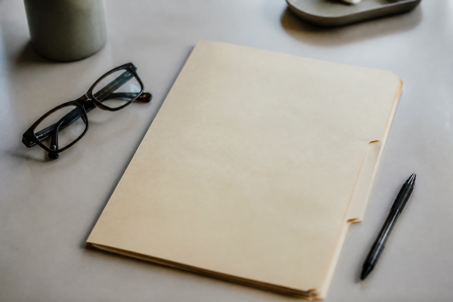 A closed buff-colored medical file folder resting on a plain desk beside a pen and a pair of reading glasses, cool daylight