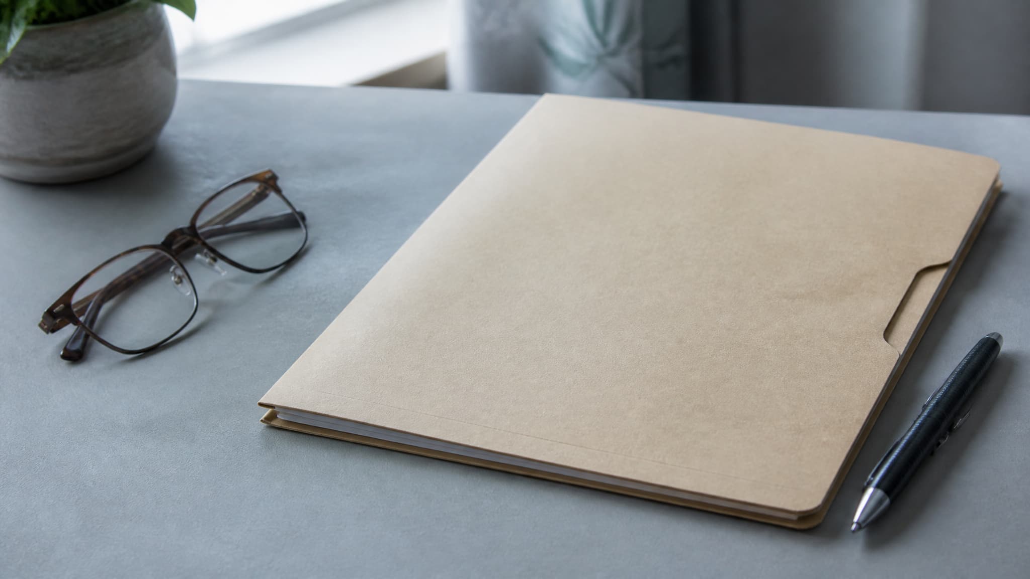 A closed medical file folder on a plain desk beside a pen and a pair of reading glasses, in cool daylight