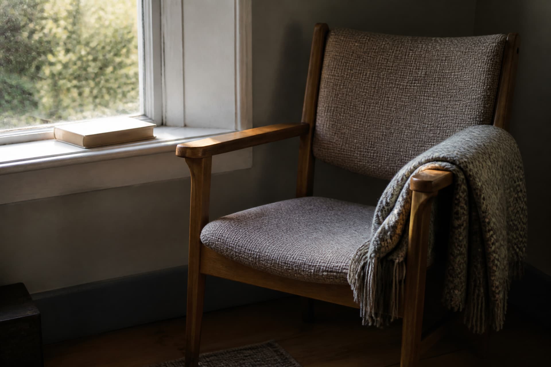 An empty chair by a window with a folded blanket over one arm and a closed book on the sill, soft late daylight