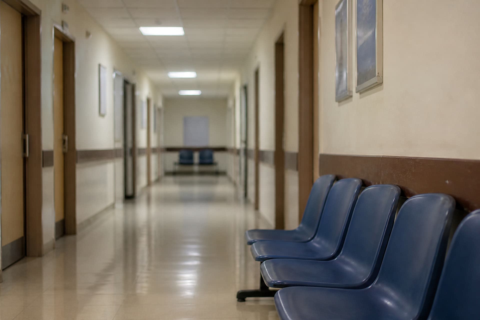 An empty hospital corridor with closed doors along one side and a row of plastic chairs against the wall, flat overhead light