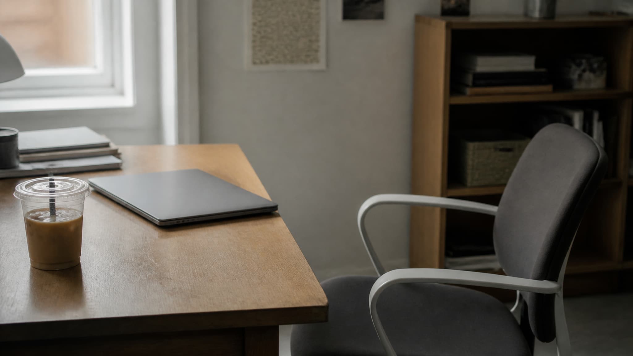 An empty office chair pushed back from a desk with a closed laptop and a cold cup of coffee, in flat morning light