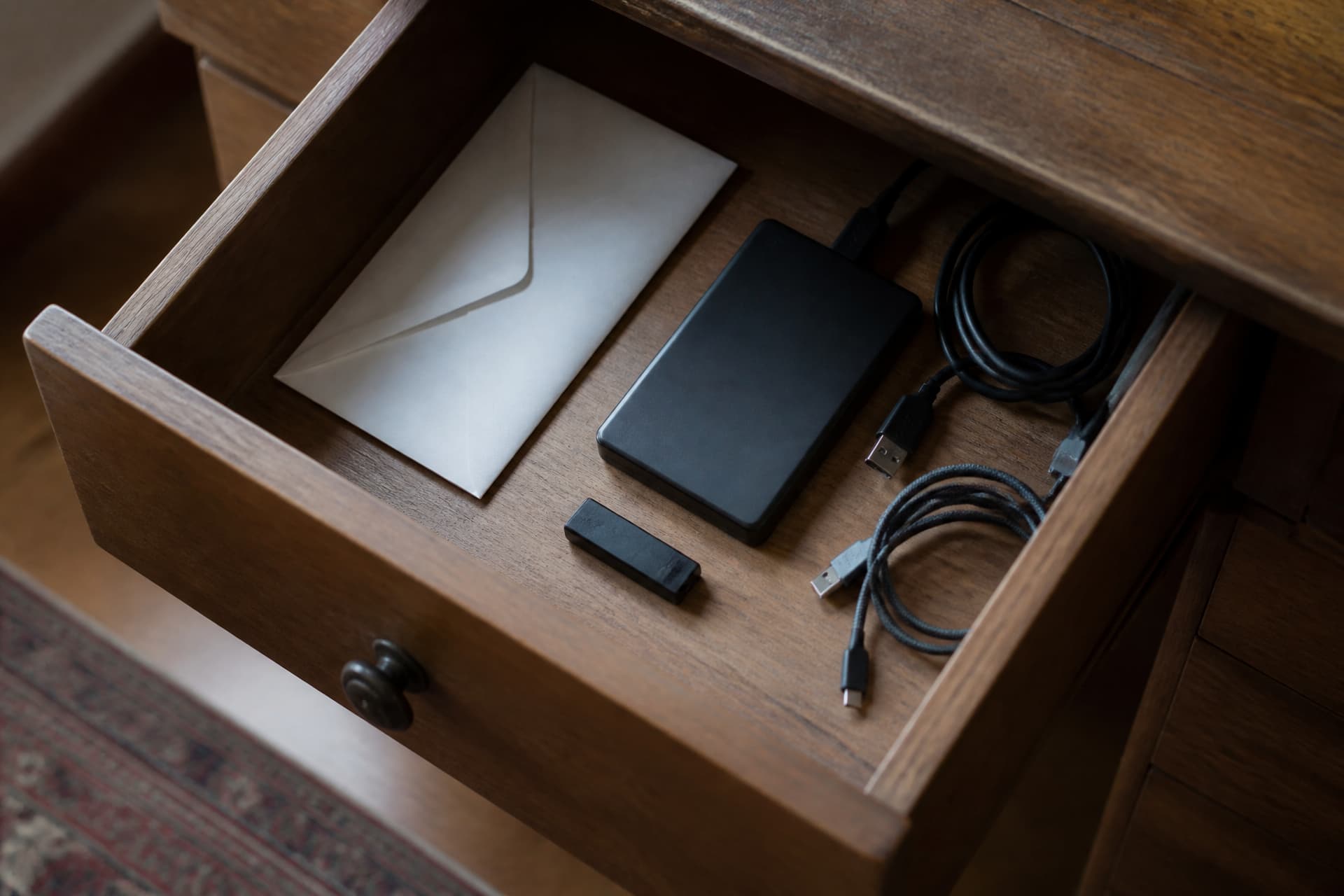 A desk drawer partly open showing a hardware wallet, a sealed envelope and an external drive among loose cables and papers