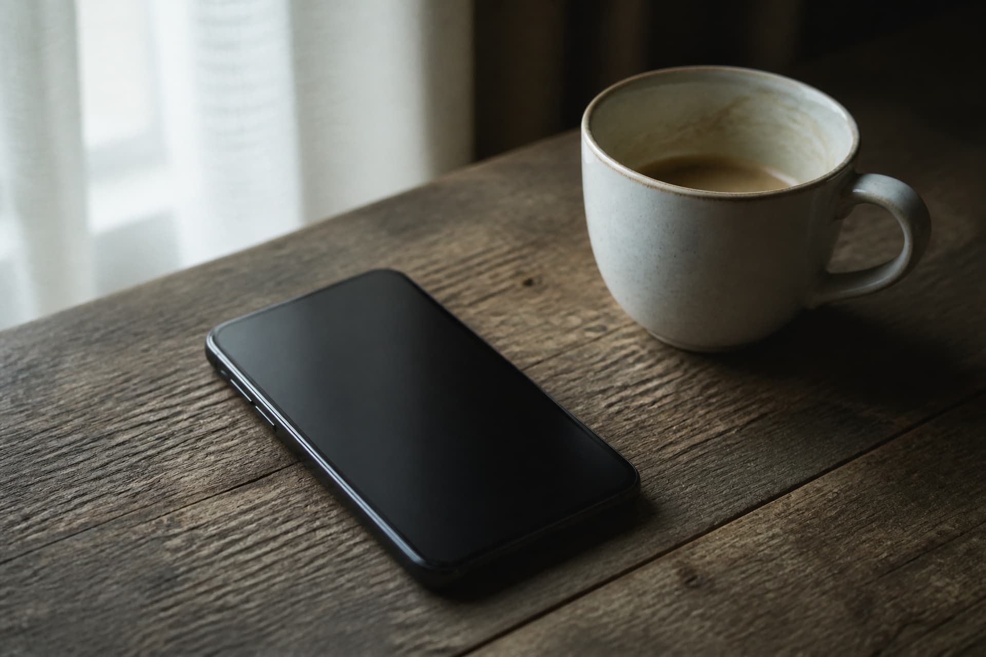 A smartphone lying face-up on a weathered wooden table with its screen completely dark, a ceramic cup of coffee beside it in soft window light