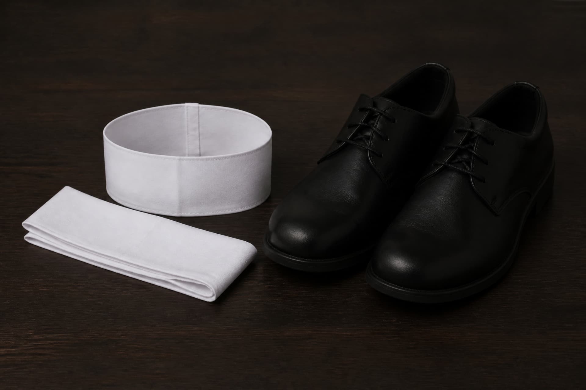 A plain white cloth armband and a folded white headband on a dark wooden table beside a pair of plain black leather shoes