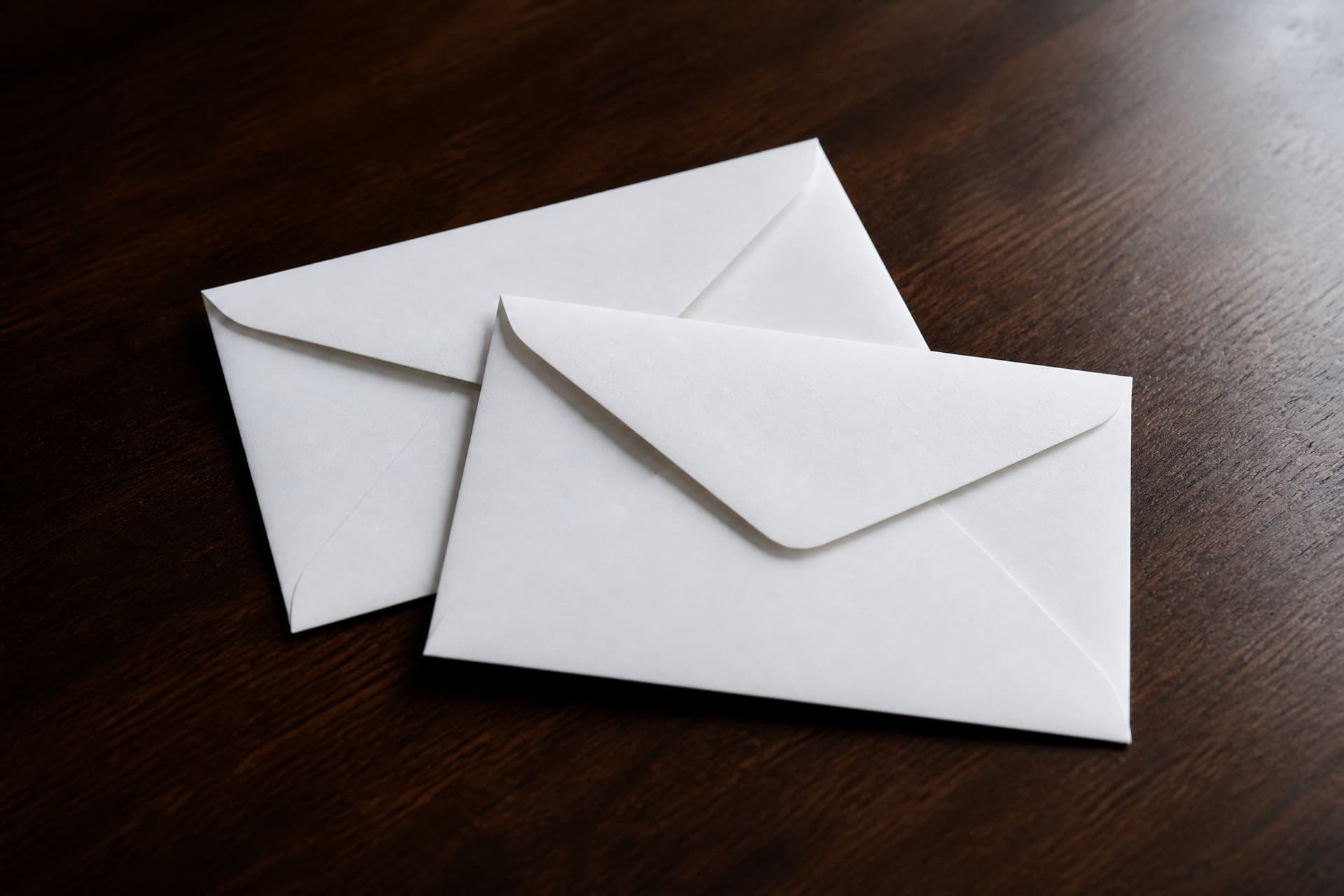 Two plain white envelopes lying on a dark wooden table in soft light