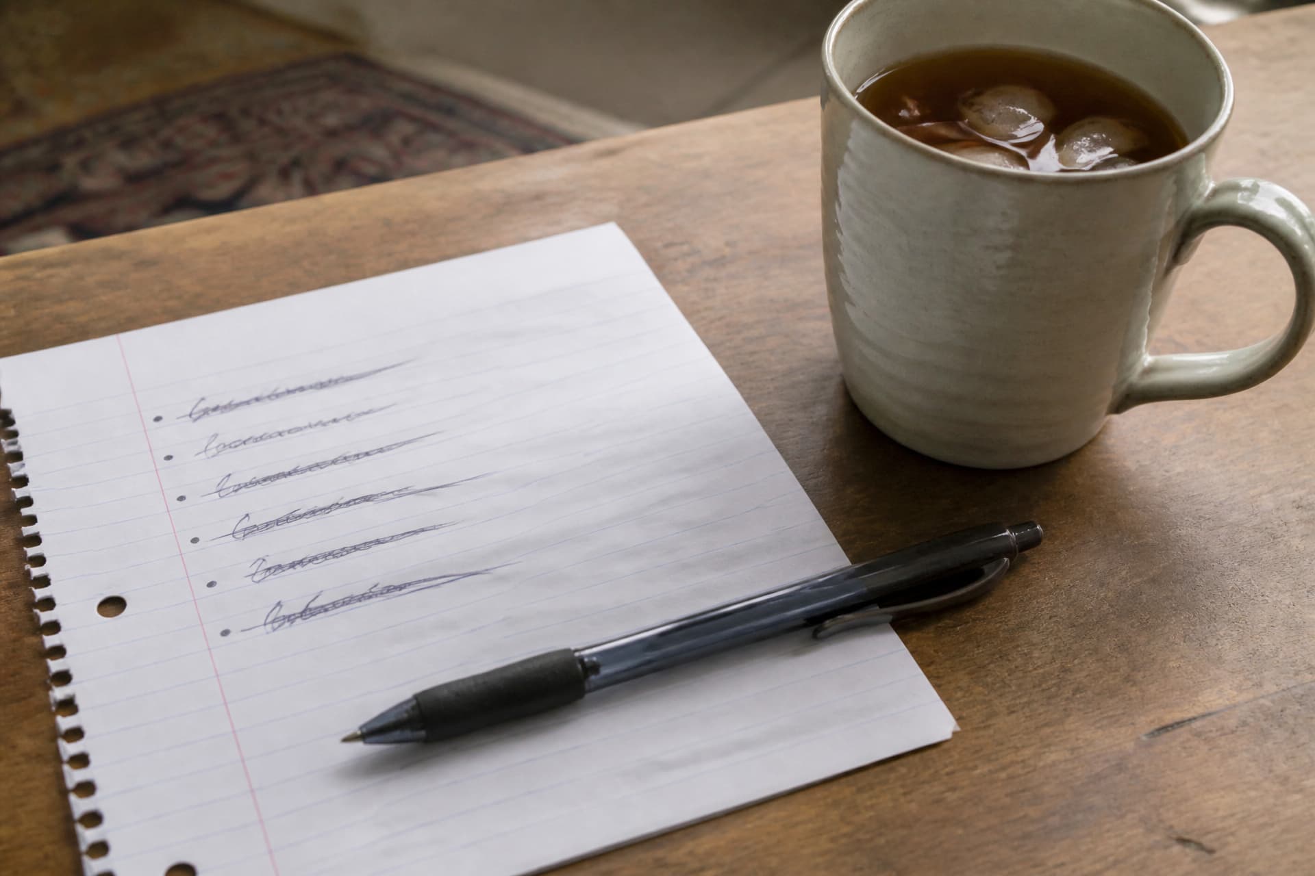 A handwritten list on lined paper with every line struck through, a ballpoint pen resting across the lower half and a pale ceramic mug beside it