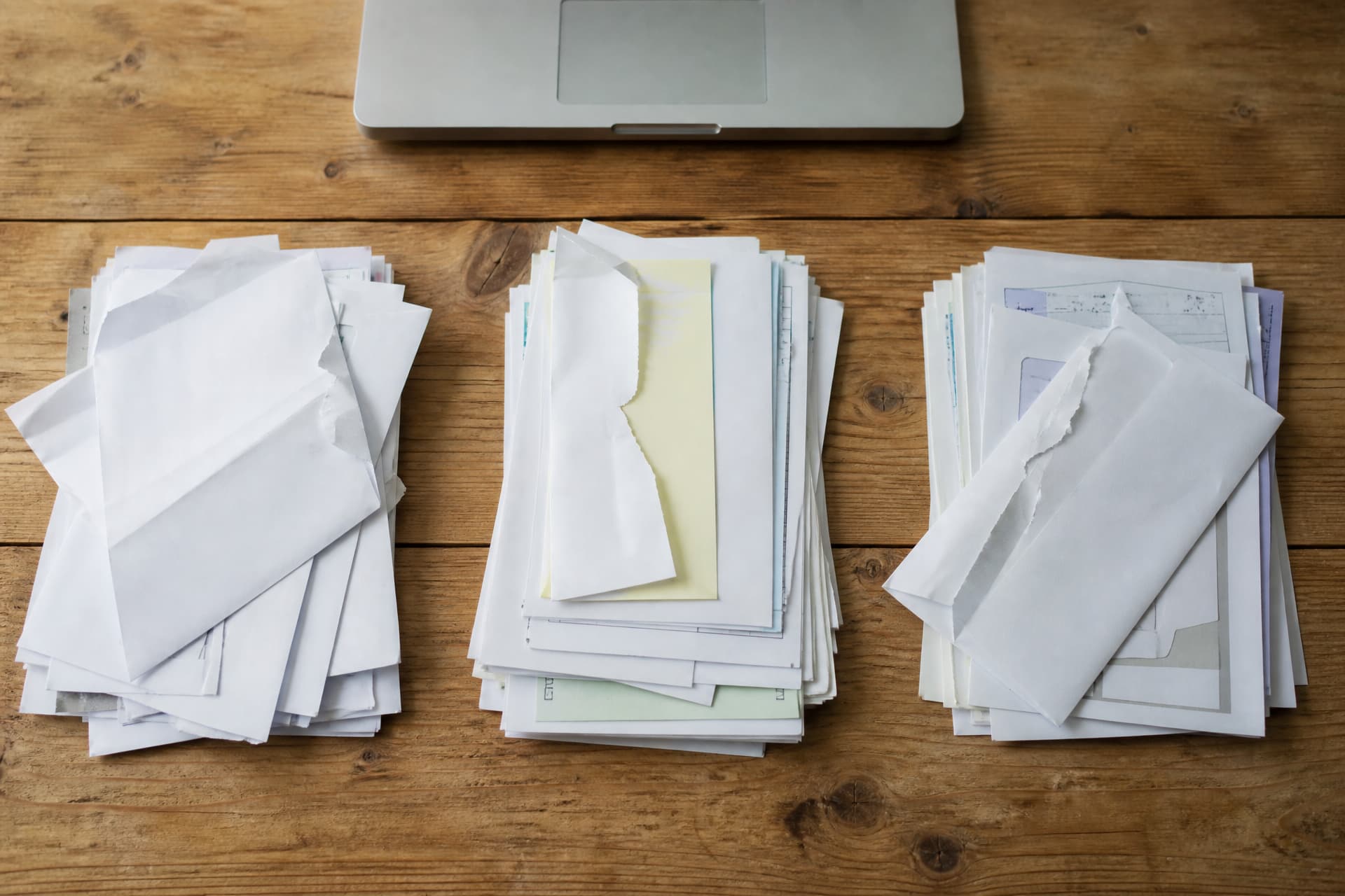 Opened mail and printed statements sorted into three separate piles on a wooden table, seen from above, with the edge of a closed laptop at the top of the frame