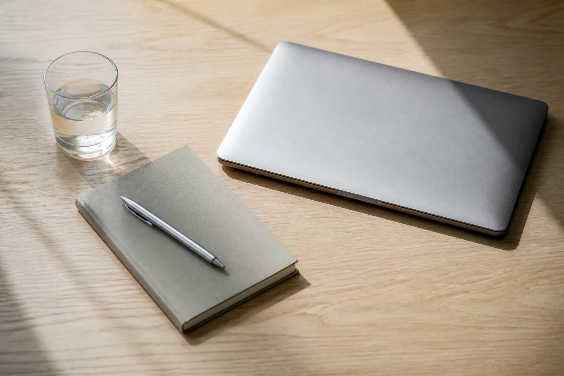 A closed silver laptop, a plain grey notebook with a pen on it and a glass of water on a pale wooden desk, seen from above in angled morning light