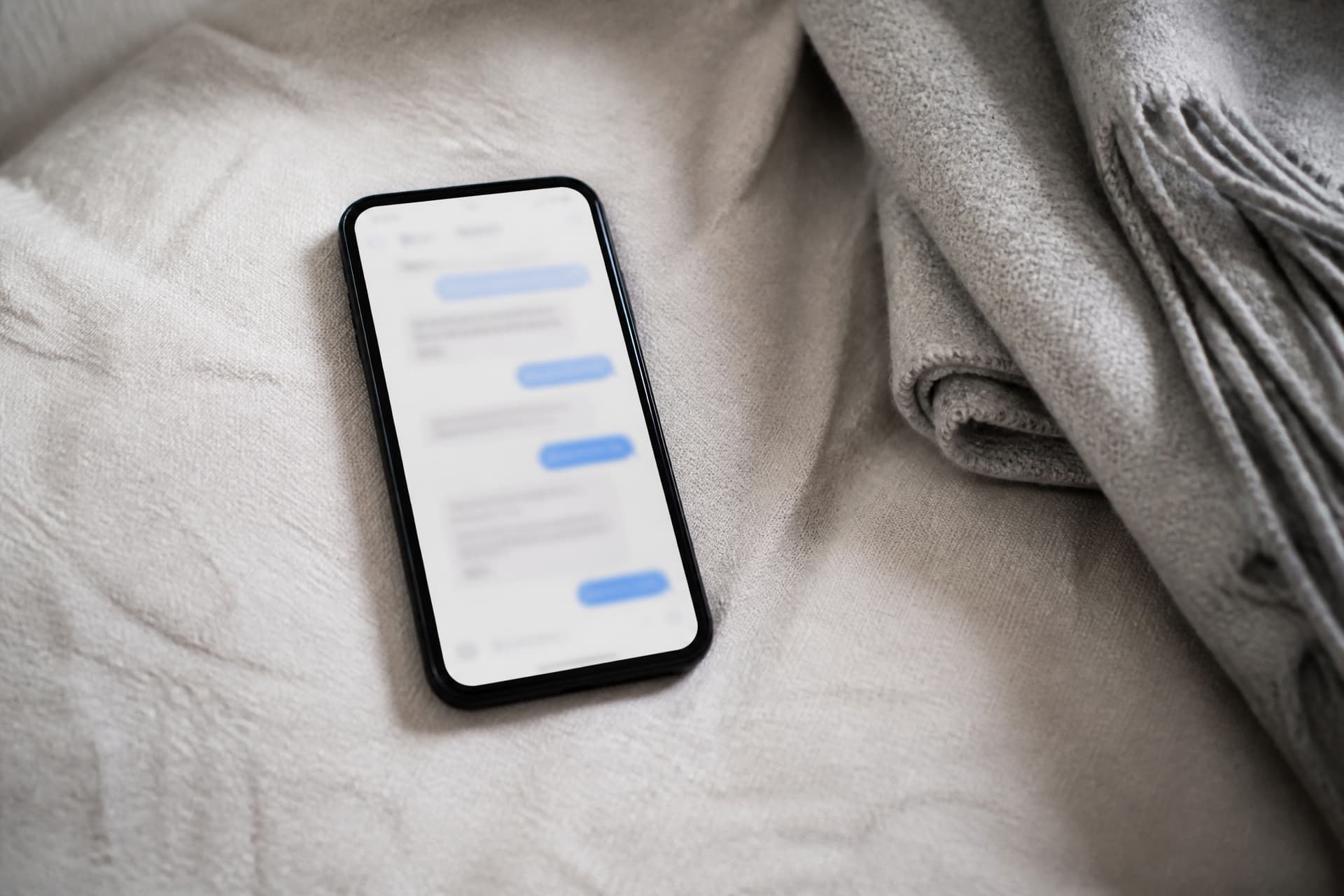 A phone lying flat on a pale linen throw seen from above, the screen showing a messaging thread of blue and grey bubbles with the words too small to read, a folded grey blanket beside it