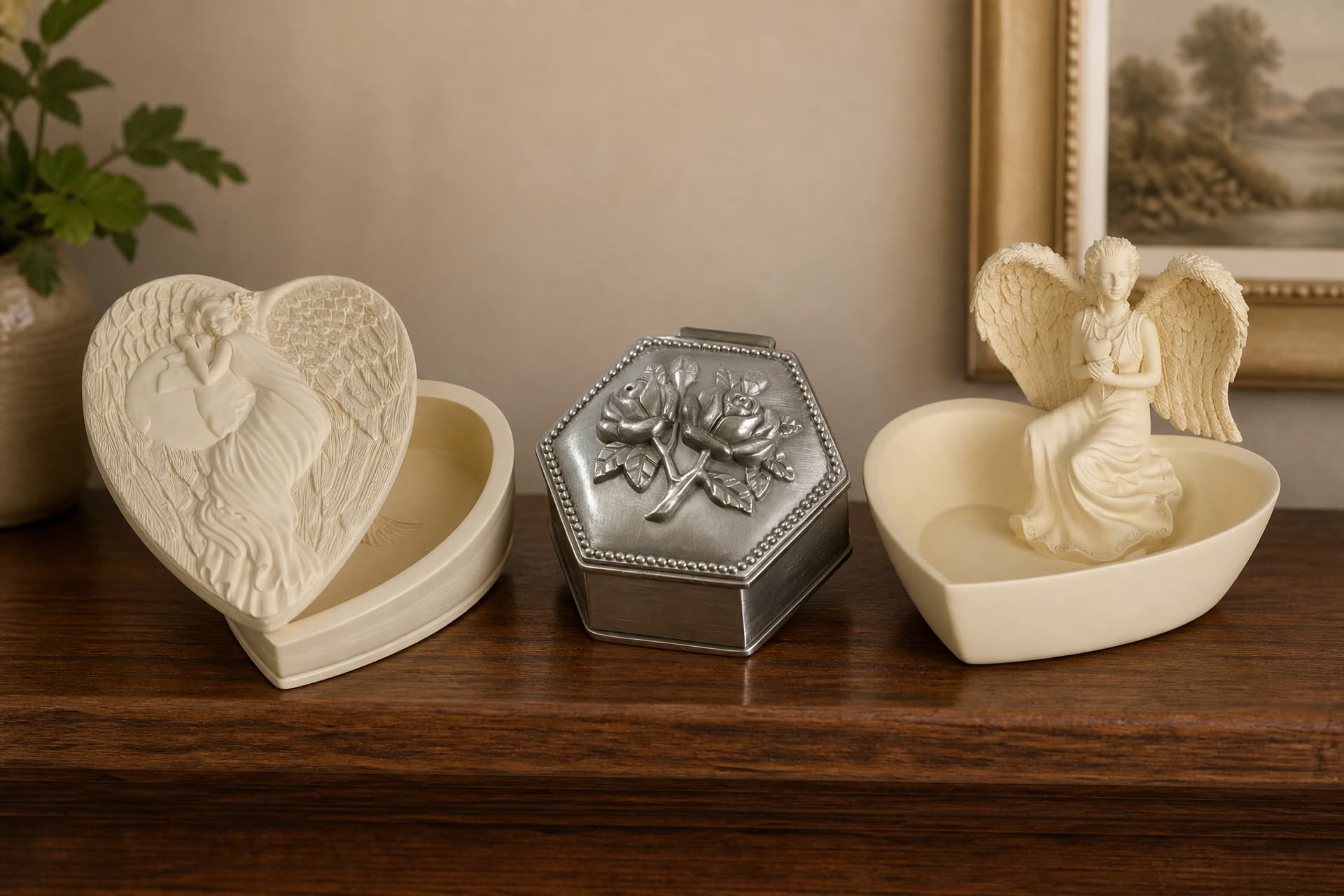 A heart-shaped cast stone keepsake box with an angel relief lid, a small pewter hexagonal box with roses, and a heart-shaped dish with a seated angel figure, on a wooden sideboard