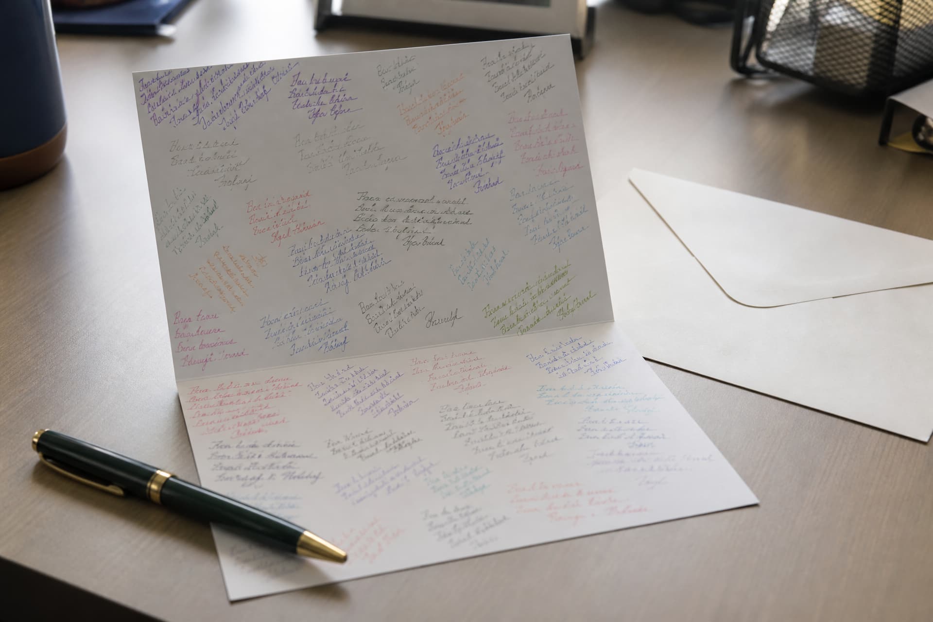 An open greeting card on a desk covered with dozens of short handwritten signatures in a dozen different pen colors, a pen and a plain envelope alongside