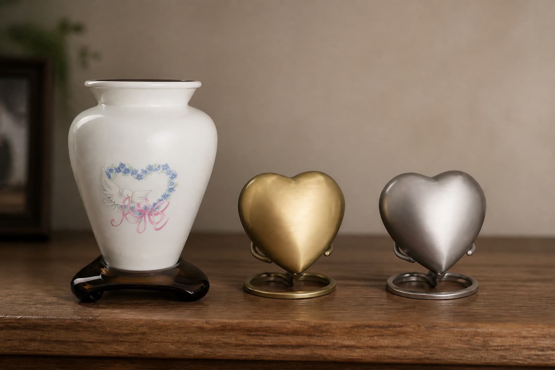 A white ceramic keepsake vase painted with a dove and a forget-me-not heart, beside a brushed gold heart keepsake and a pewter heart keepsake, each on a small ring stand