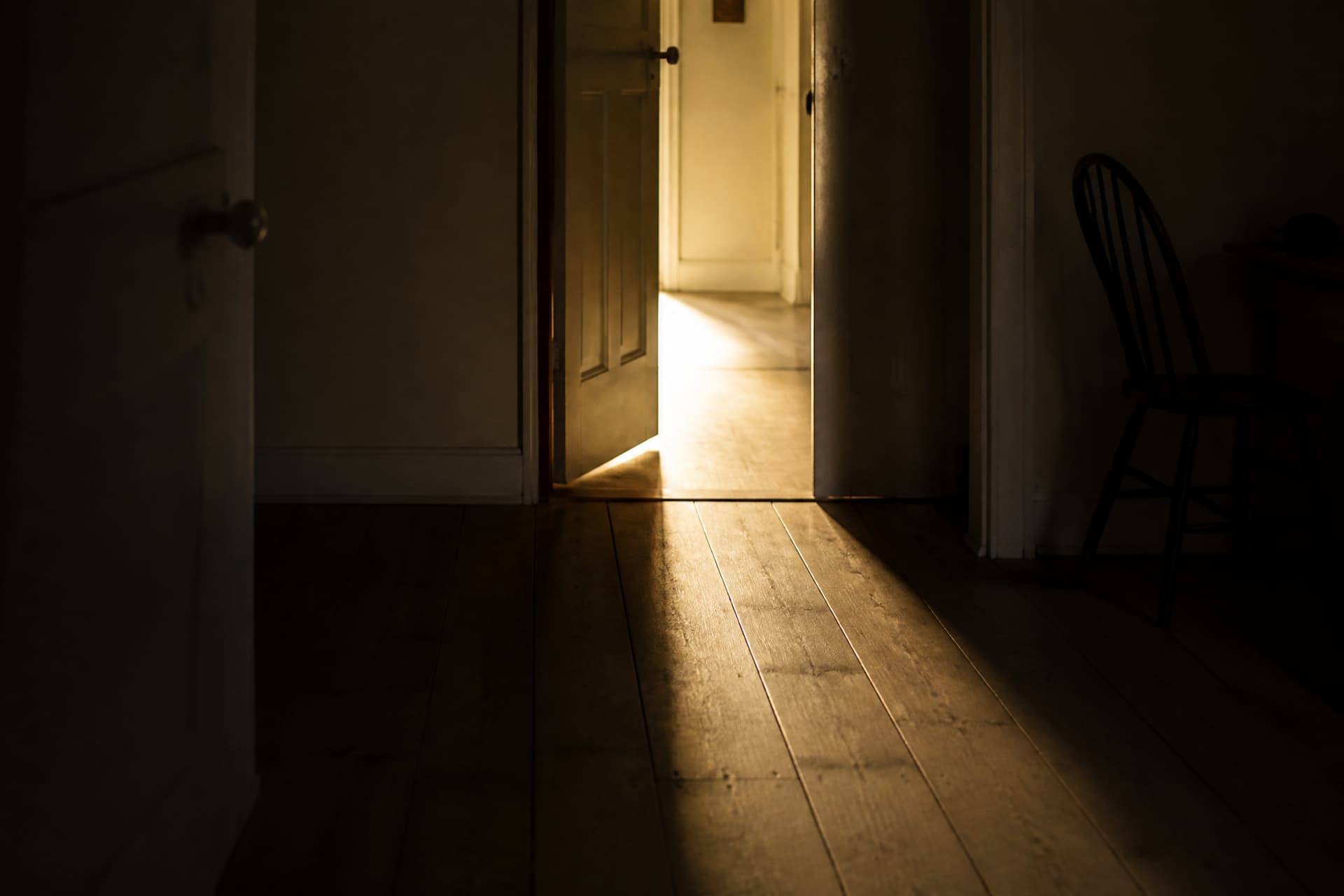 A doorway seen from a dark room, warm light spilling across the floor from the lit hallway beyond, nobody in the frame