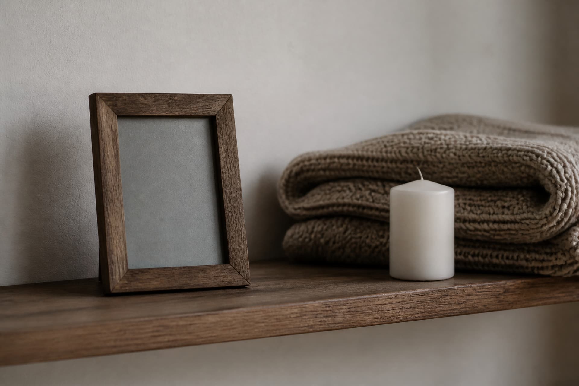 A narrow wooden shelf holding an empty wooden picture frame with plain grey card in it, a folded knitted blanket and an unlit white candle