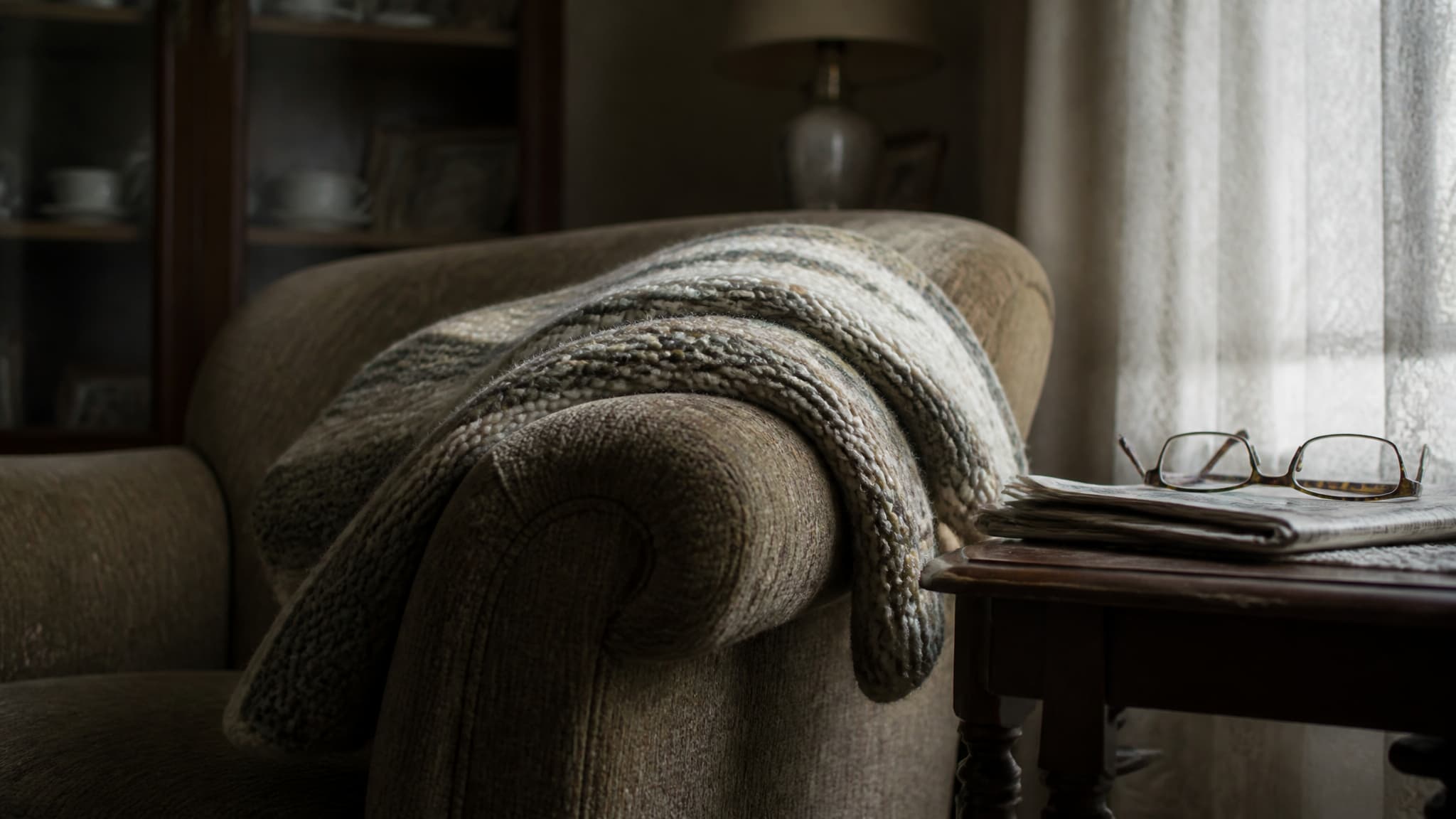 A knitted wool blanket draped over the arm of a worn armchair in a dim living room, reading glasses resting on a folded newspaper on the side table beside it