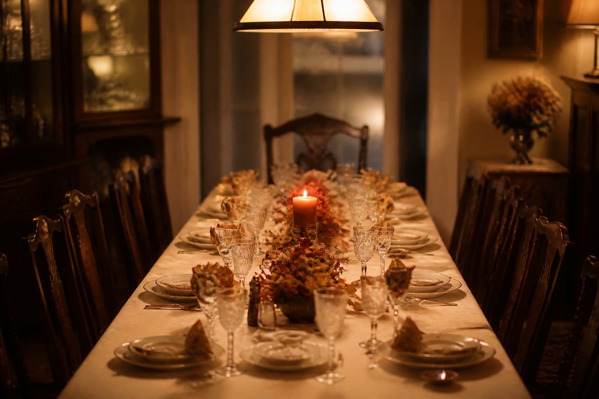A long dining table laid for a holiday meal with china, cut glass and a lit candle, the carved chair at the head of the table empty and pushed back