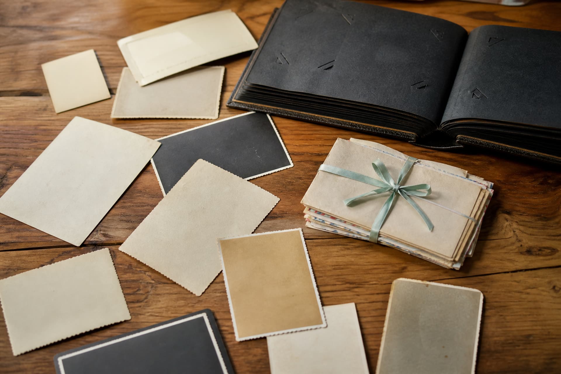 Old photographs spread face down across a wooden table beside an open black-paged album and a bundle of handwritten letters tied with a ribbon
