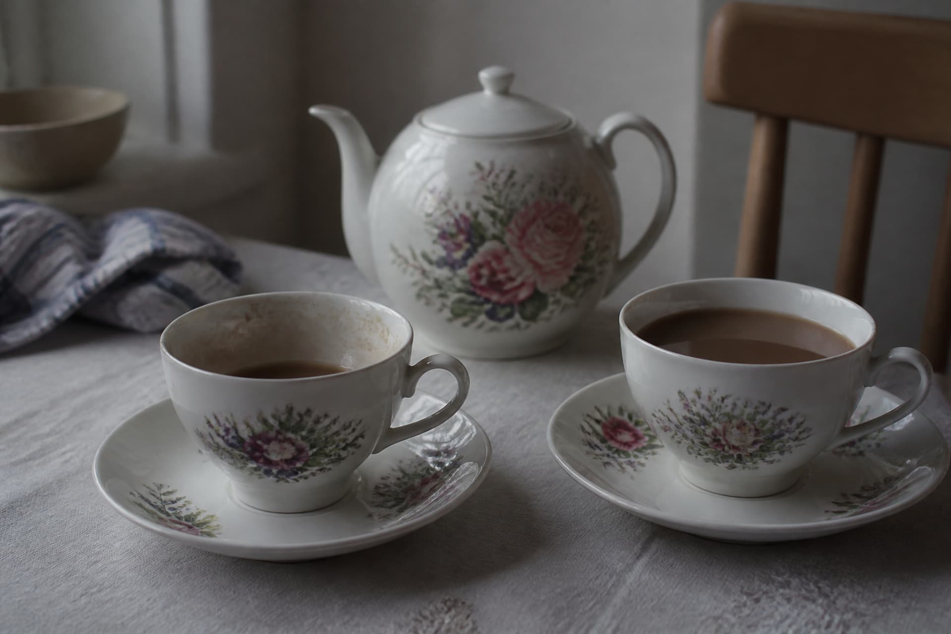Two china teacups on saucers on a kitchen table, one half drunk and one untouched, a floral teapot behind them in flat afternoon light