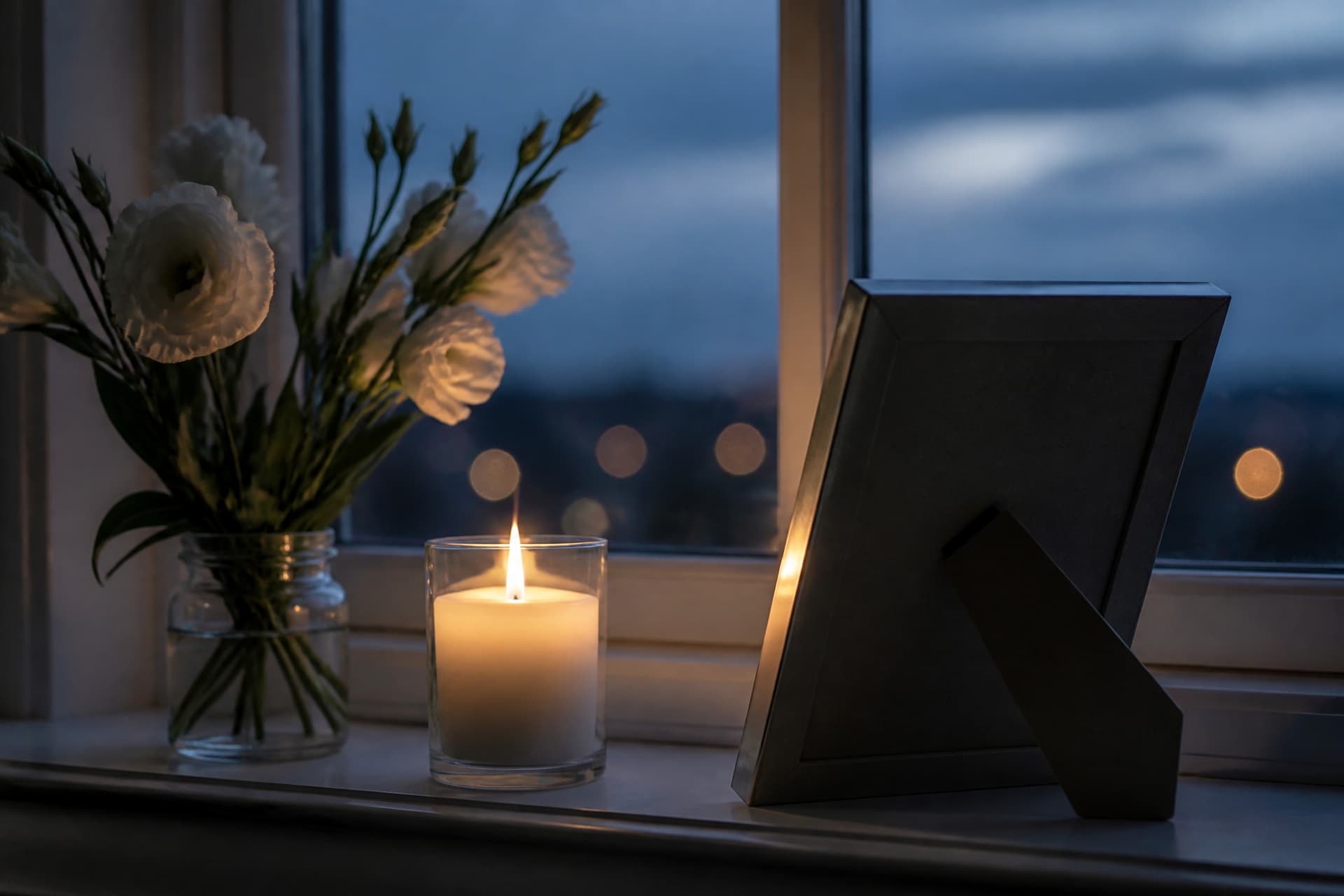 A single lit memorial candle on a windowsill beside a framed photograph turned slightly away from the camera