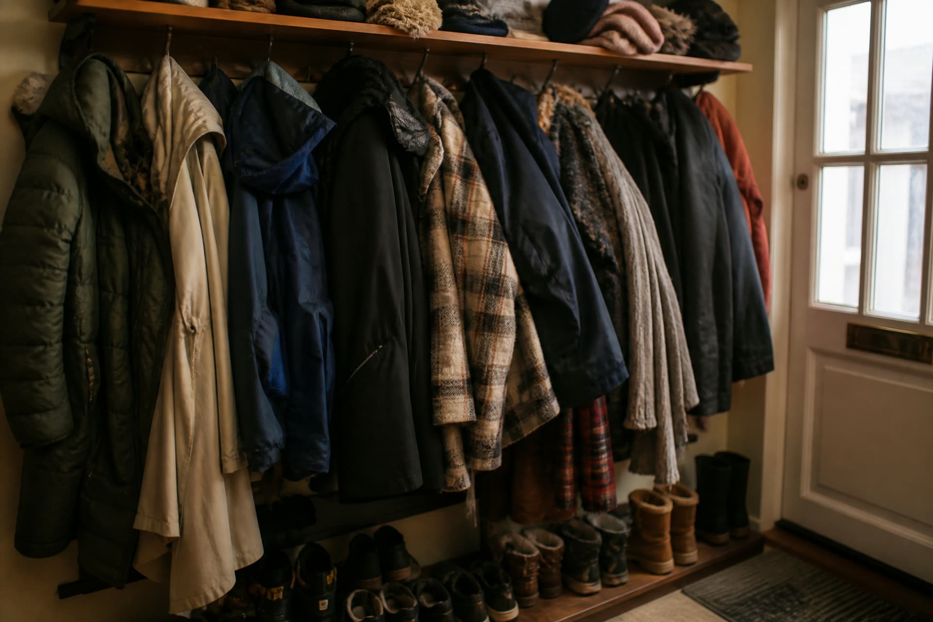 A crowded coat rack by a front door with a dozen coats and scarves hung over each other, shoes lined up beneath, a hallway light on