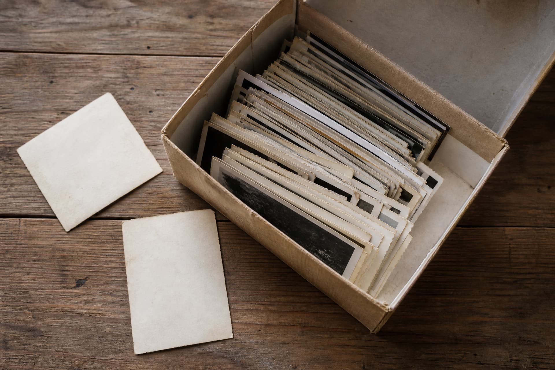 An open cardboard shoebox of loose old photographic prints on a bare wooden table, with two prints lying face down beside it showing only their blank backs
