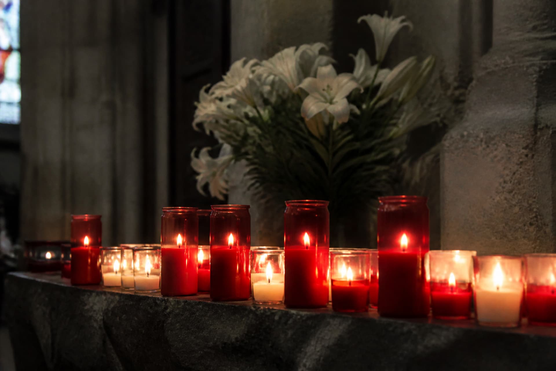 A row of lit glass votive candles on a stone ledge in a dim church interior, with white flowers behind them