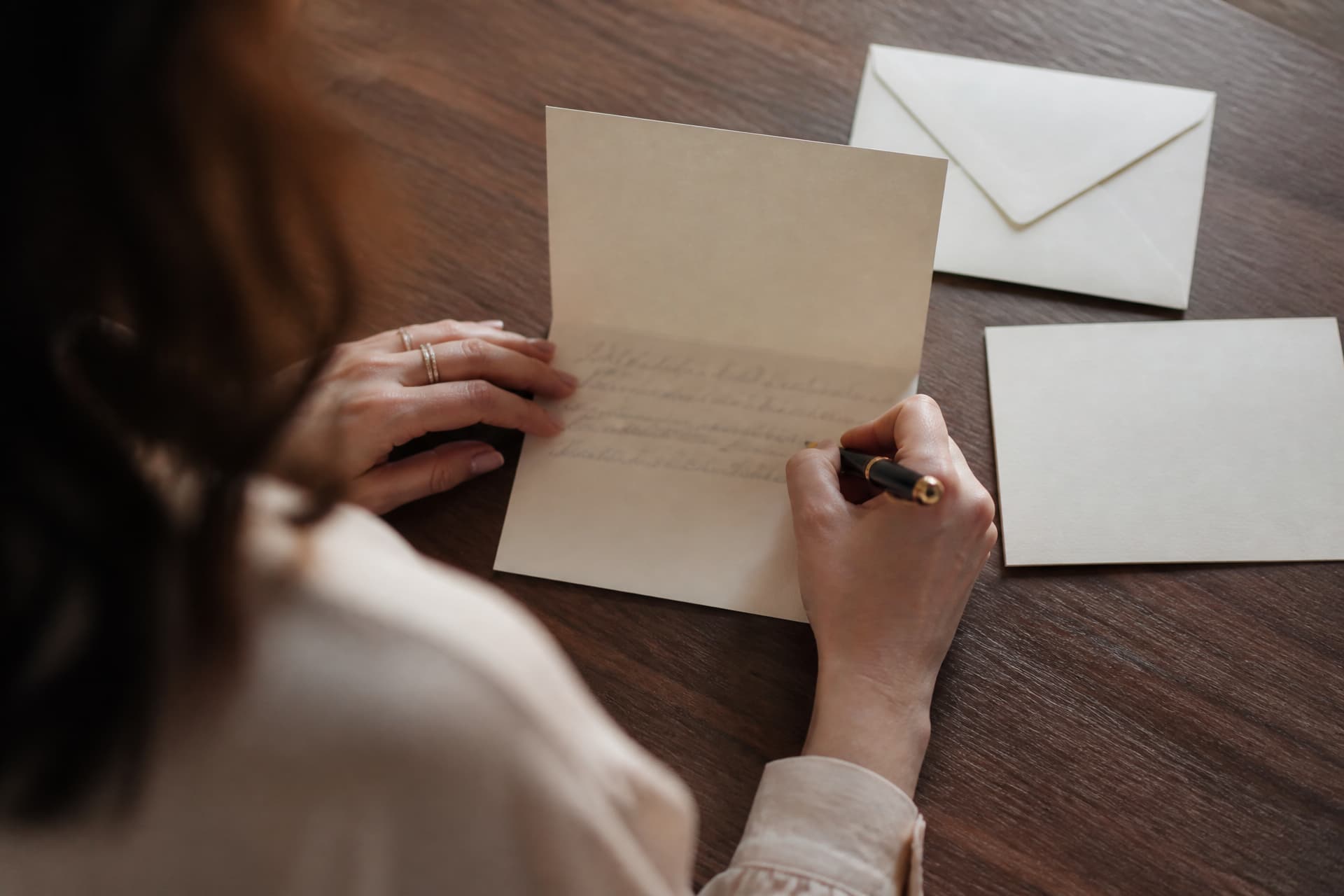 A woman's hands writing with a fountain pen inside a folded cream card at a dark wooden table, seen from over the shoulder, with a plain envelope and a second card alongside