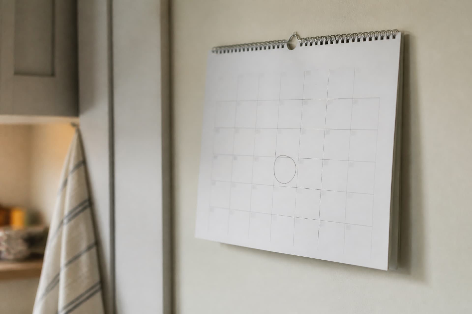 A wall calendar hanging in a quiet kitchen with a single date circled in pencil, seen at an angle in soft afternoon light