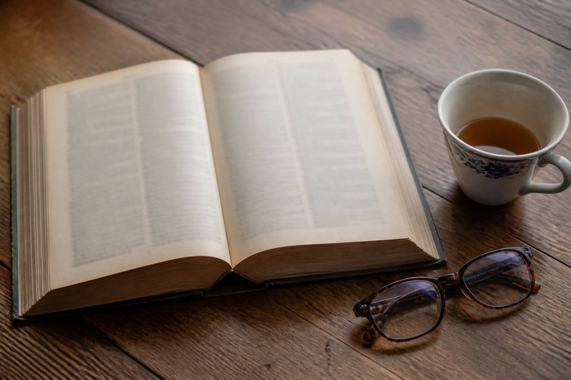 An open hardback dictionary on a dark wooden table beside a pair of tortoiseshell reading glasses and a half-finished cup of tea, its pages out of focus