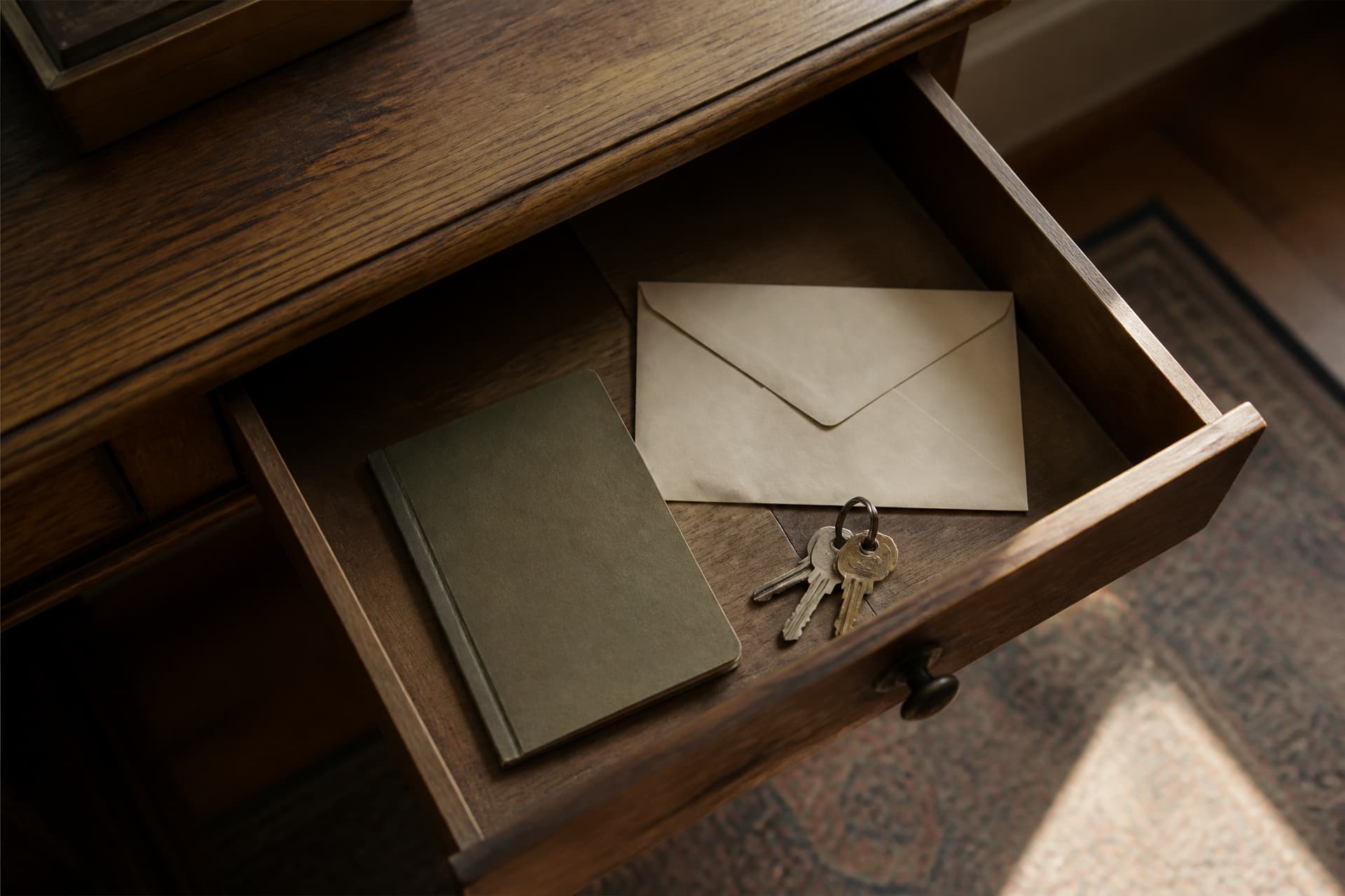 An open desk drawer holding a sealed envelope, a small notebook and a set of keys, seen from above in warm light