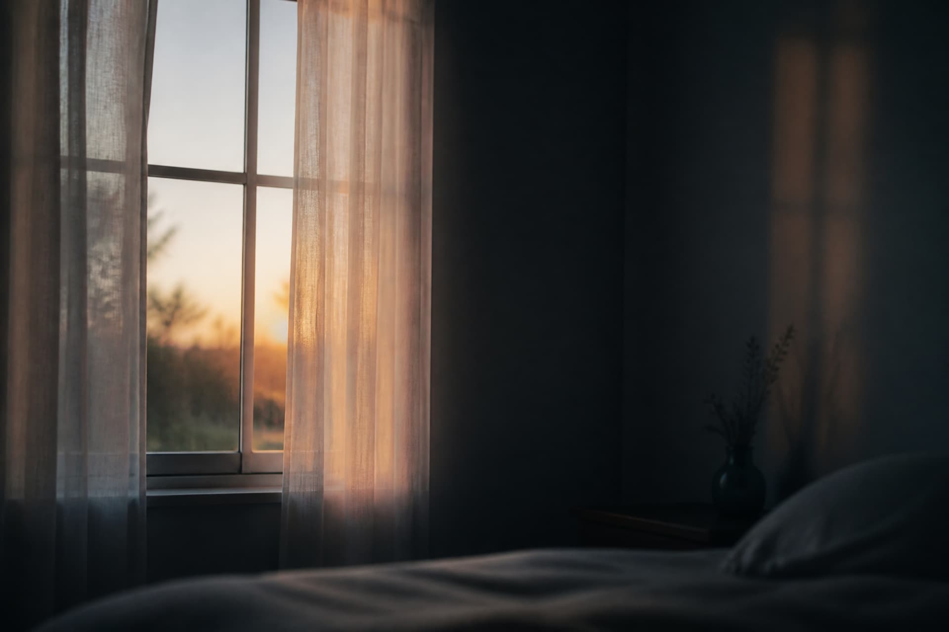 The first pale light of dawn at a curtained window, seen from inside a dim bedroom