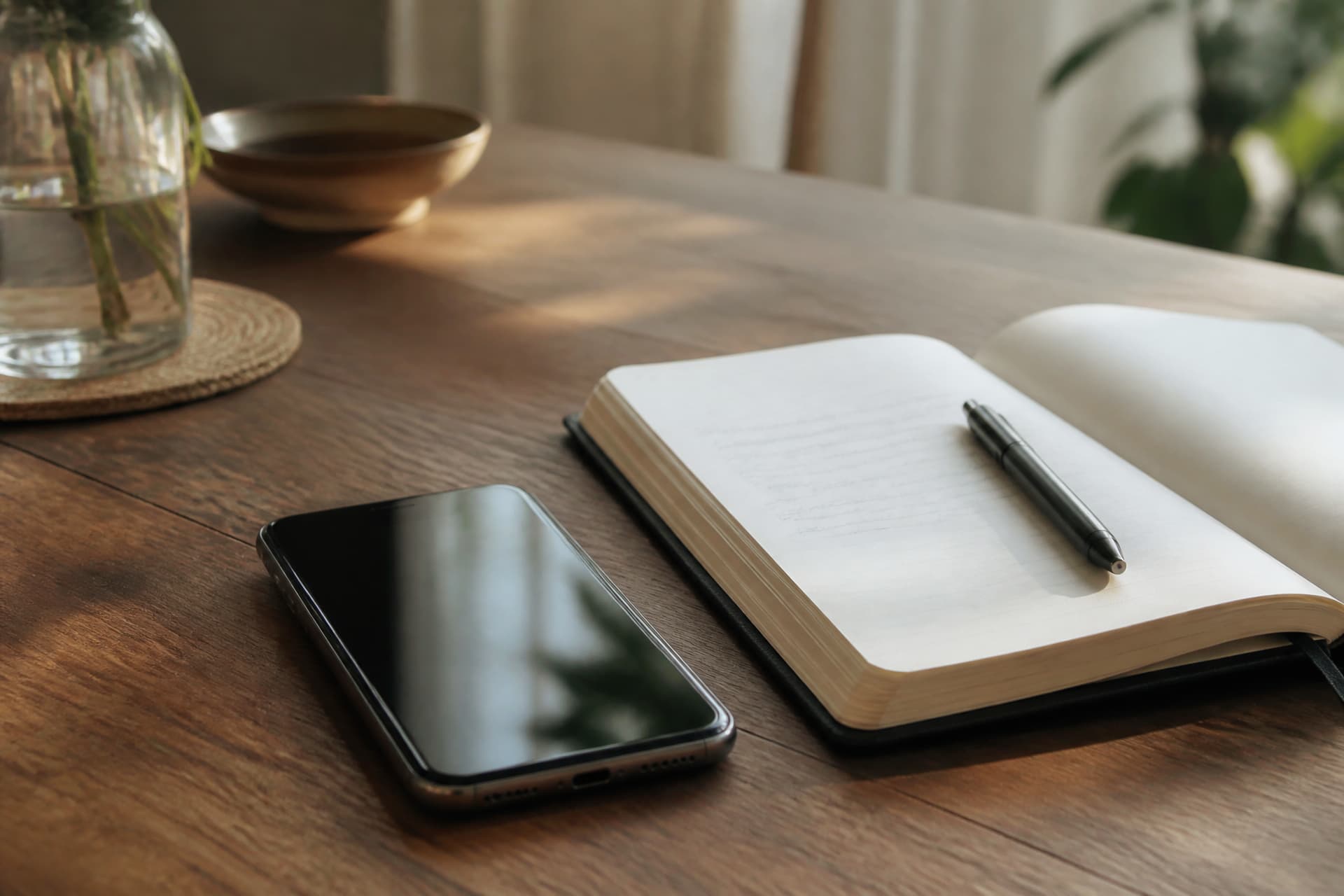 A phone lying face-up beside an open notepad with a short handwritten list and a pen, on a wooden table in afternoon light