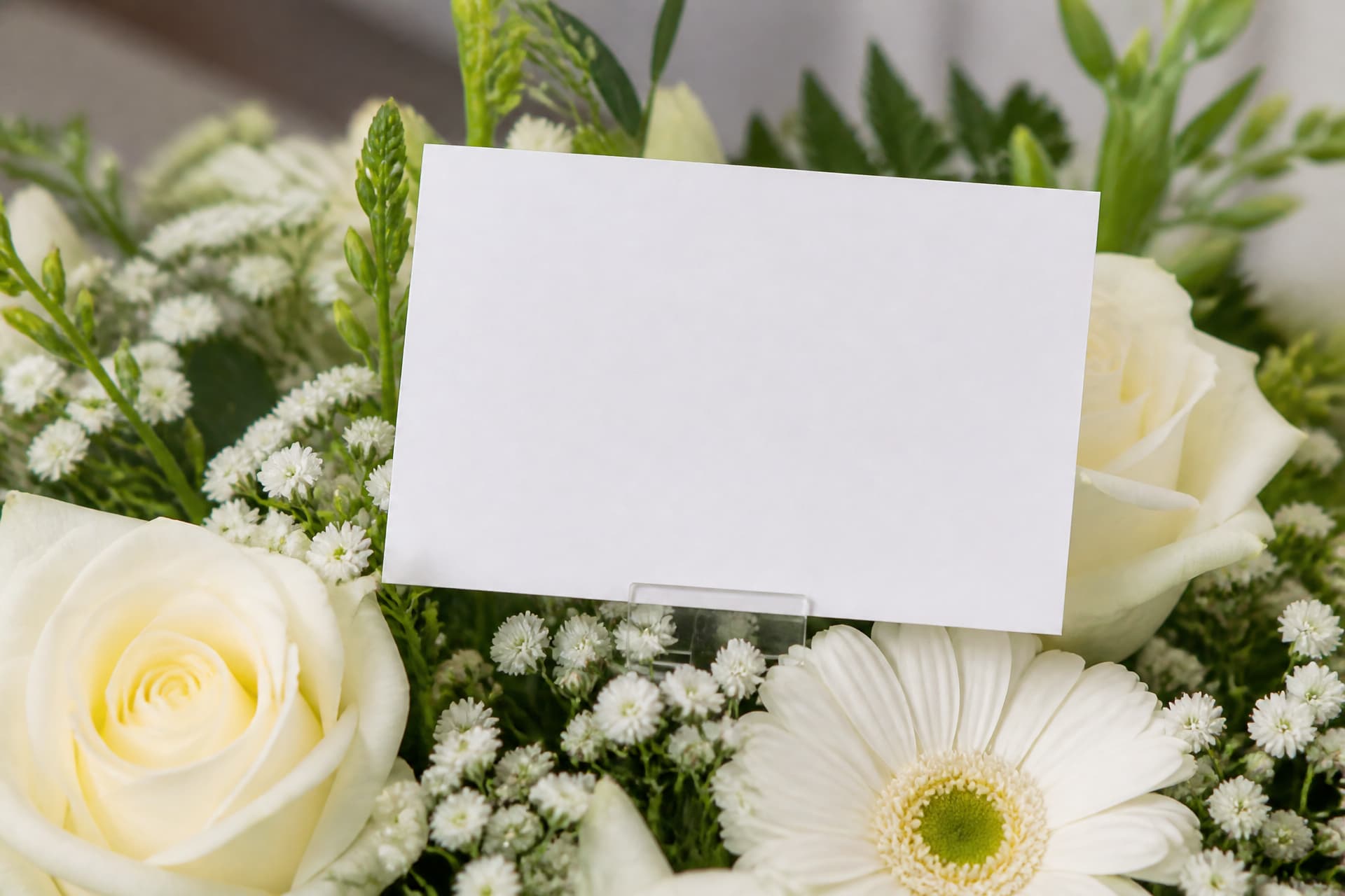 A blank white florist's enclosure card on a clear plastic pick, tucked among white roses, gerberas and gypsophila