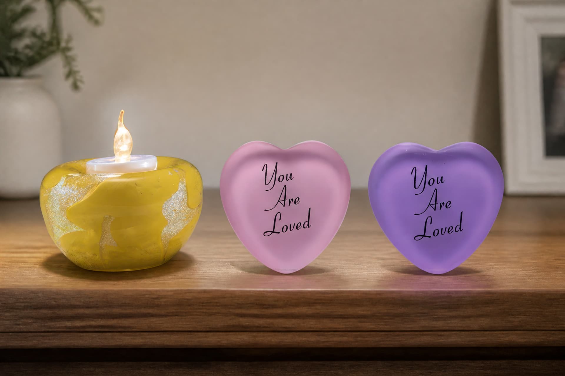 A yellow swirled glass tealight holder with a lit candle beside two frosted glass hearts, one pink and one purple, each engraved "You Are Loved"