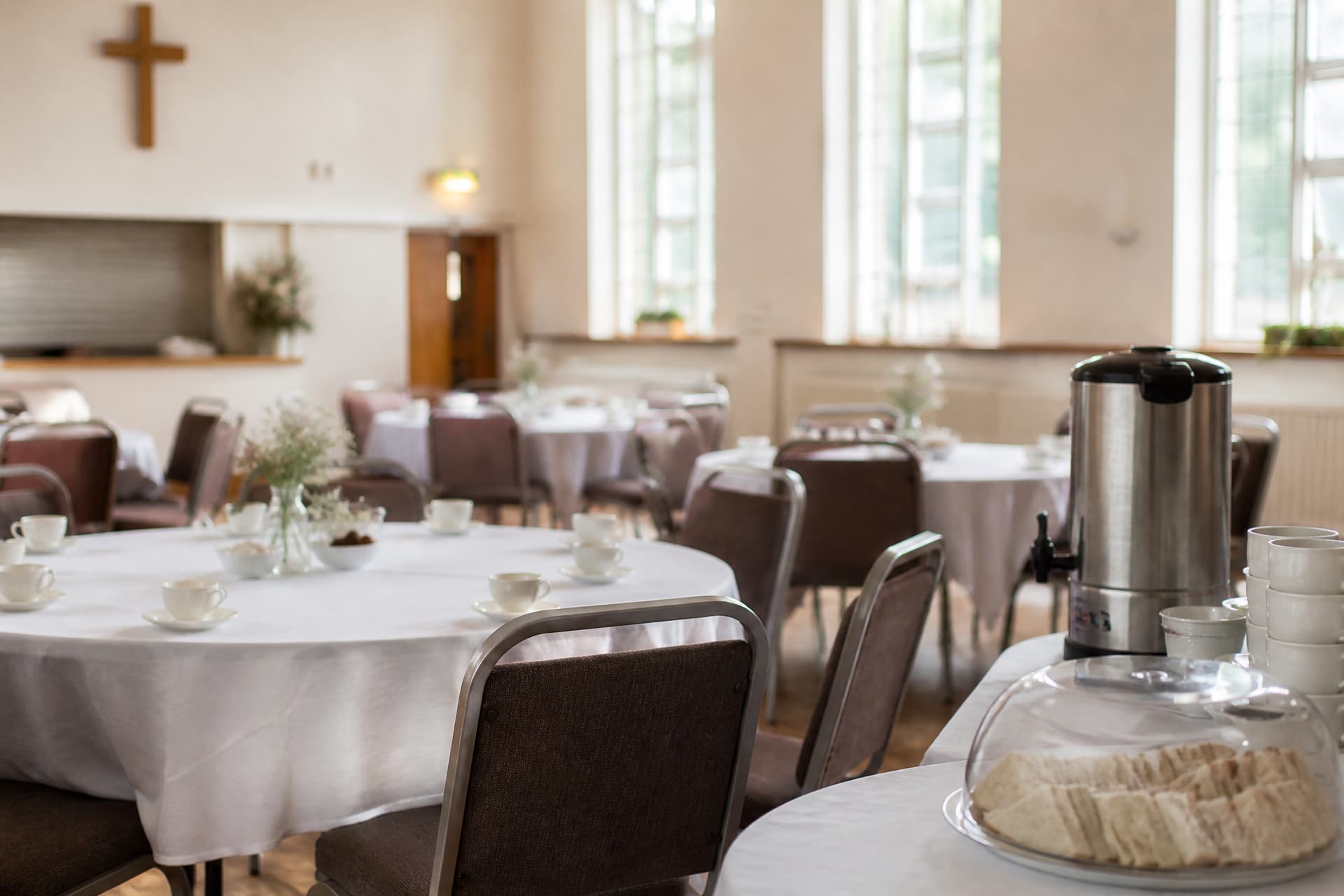 A church hall set for a reception after a service, round tables with white cloths, cups and saucers, and a tea urn on a side table
