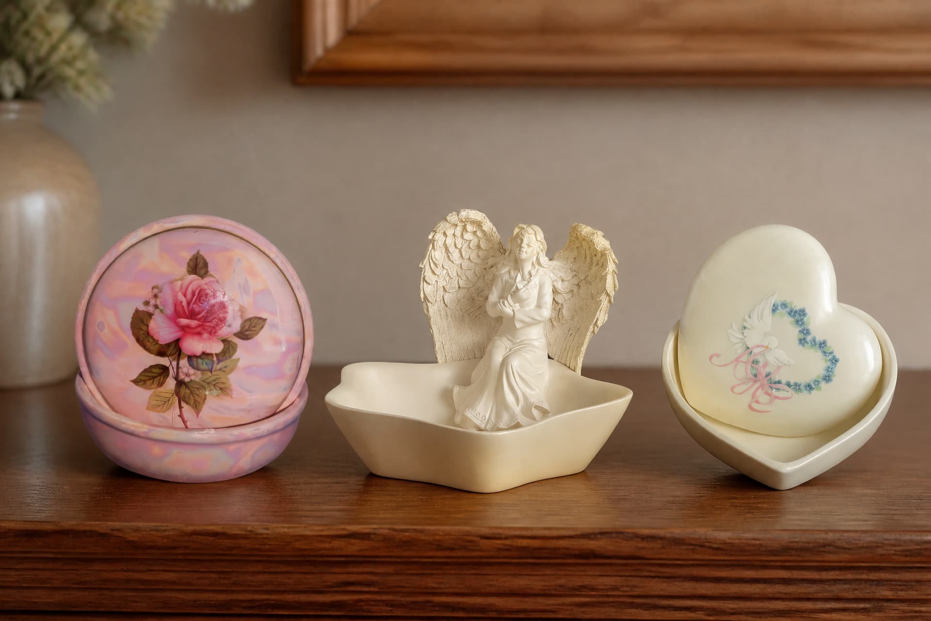 Three small ceramic keepsake boxes on a wooden shelf, one round with a painted rose, one holding a sculpted angel figure, and one heart-shaped with doves