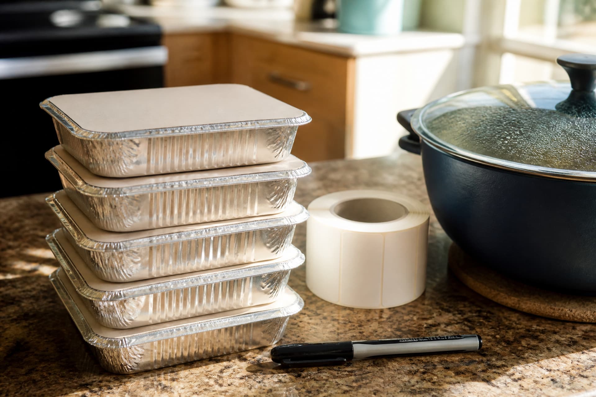 A stack of five clean foil containers with card lids on a kitchen counter, a roll of blank labels and a black marker beside them, a covered pot cooling at the edge of the frame