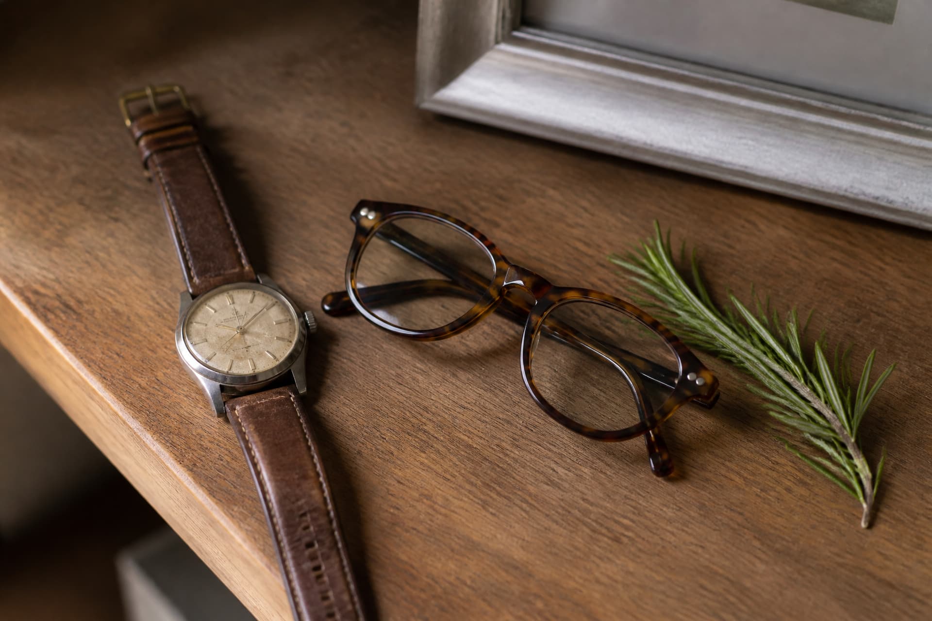 A close view of a shelf top showing a worn leather watch, a pair of reading glasses and a small sprig of rosemary beside a photo frame edge