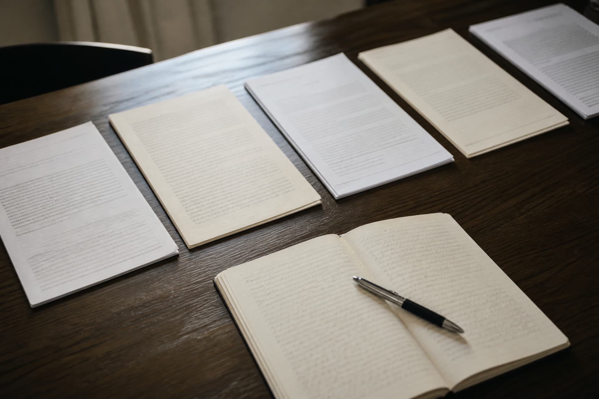 Several certified documents laid out in a row on a desk beside a notebook listing institution names, a pen across the notebook