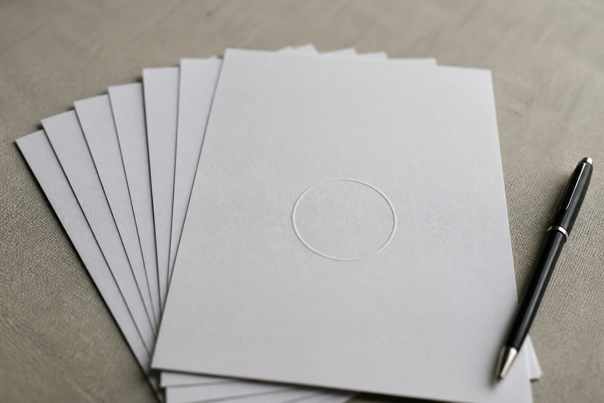 A small stack of printed documents fanned slightly on a plain table, the topmost carrying a blank embossed circular impression, beside a pen